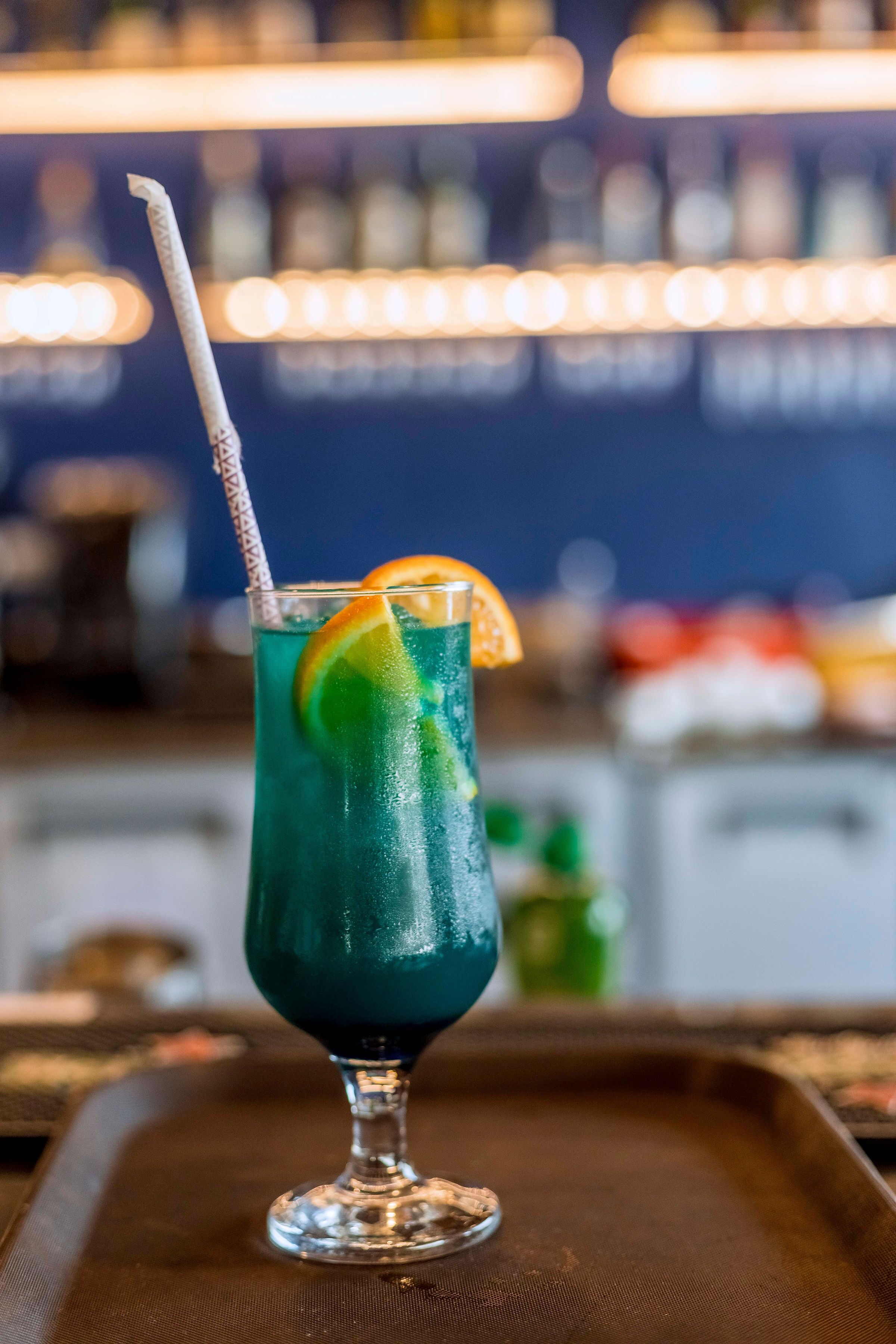 Ein hoher Glas mit Trinkhalm, gefüllt mit einem blauen Getränk, garniert mit einer Orangenscheibe, auf einem Tablett.