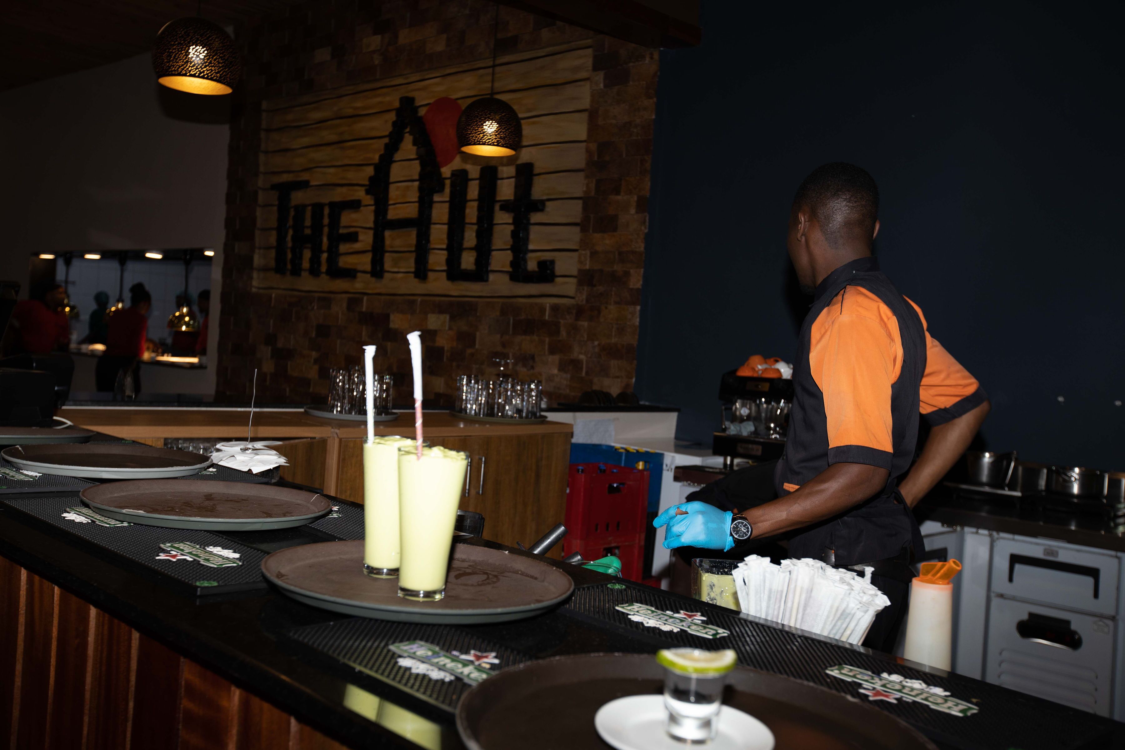 Eine Bar mit Holzwand und hängenden Lampen, ein Barkeeper in Uniform sowie verschiedene Barutensilien wie Gläser und Teller.