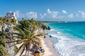 A coastal scene with a clear view of turquoise waters, a sandy beach, and a stone structure on a cliff.