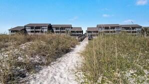 Um hotel na praia com vários edifícios, um caminho de areia que leva à entrada e um céu azul límpido.