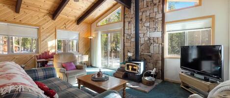 A cozy cabin interior with a stone fireplace, wooden beams, plaid furniture, and a television.