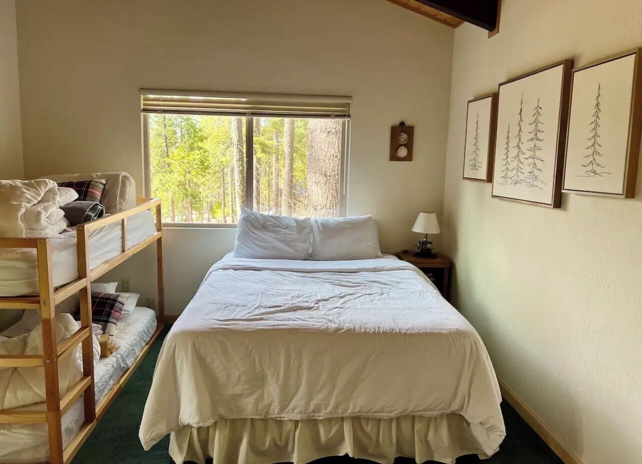 Quarto com cama de solteiro, janela com vista para árvores e quadros na parede.