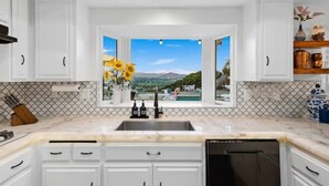 A kitchen with white cabinets, a marble countertop, a black dishwasher, and a view of a pool and mountains through a window. - Central Location, Spectacular Views, Pool, Hottub (Escondido)
