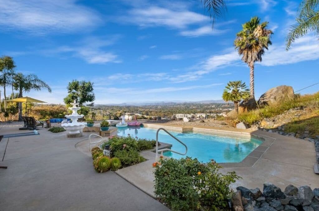 A swimming pool with a fountain, surrounded by greenery and palm trees, with a clear view of the landscape in the background.