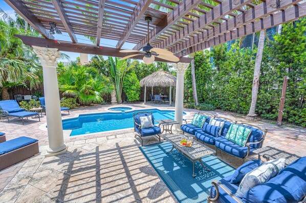 A pool area with a covered seating area, a thatched-roof structure, and lush greenery.