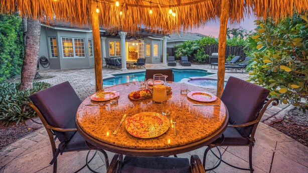 An outdoor dining area with a thatched roof, a pool, and a patio with chairs and a table set for a meal. - Steps from the Beach / Heated Pool / Seaside Key (Fort Lauderdale)