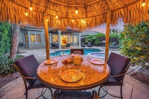 An outdoor dining area with a thatched roof, a pool, and a patio with chairs and a table set for a meal.