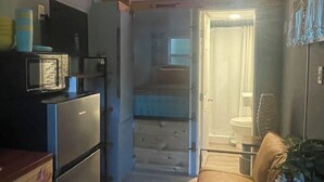 Une cuisine compacte équipée d’un four à micro-ondes, d’un réfrigérateur et de meubles en bois. On aperçoit un petit coin repas avec un canapé et une plante.