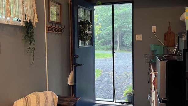 Une pièce chaleureuse avec un canapé marron, une décoration murale en macramé, un paillasson et une vue sur une zone boisée, visible à travers la porte.