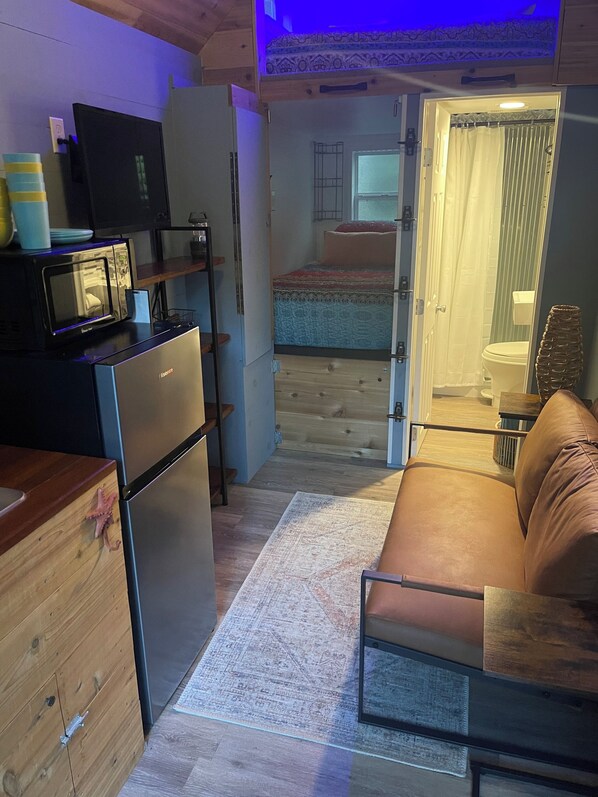 Uma cozinha compacta com geladeira, micro-ondas e uma pequena mesa de jantar. Um sofá está localizado na área de estar, e um piso de madeira é visível.