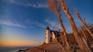 A lighthouse with a white exterior, a red roof, and a black top, situated next to a body of water with a clear sky.