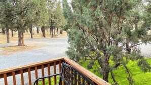 Une terrasse en bois, dotée d’une rambarde métallique, surplombe un chemin de gravier et une zone herbeuse parsemée d’arbres.