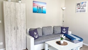 Ein modernes Wohnzimmer mit einem weißen Sofa, grauen Kissen, einem weißen Couchtisch und einem blauen Teppich. Auf einem Holzschrank stehen Blumentöpfe, an der Wand hängt ein Gemälde und es steht eine Stehlampe da.