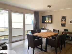 Een eethoek met een houten tafel en stoelen, een wandtelevisie en een raam met zonwering.