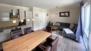 Eine moderne Küche mit Esstisch und Stühlen, ein Wohnbereich mit Sofa und Couchtisch sowie ein großes Fenster mit Vorhängen.