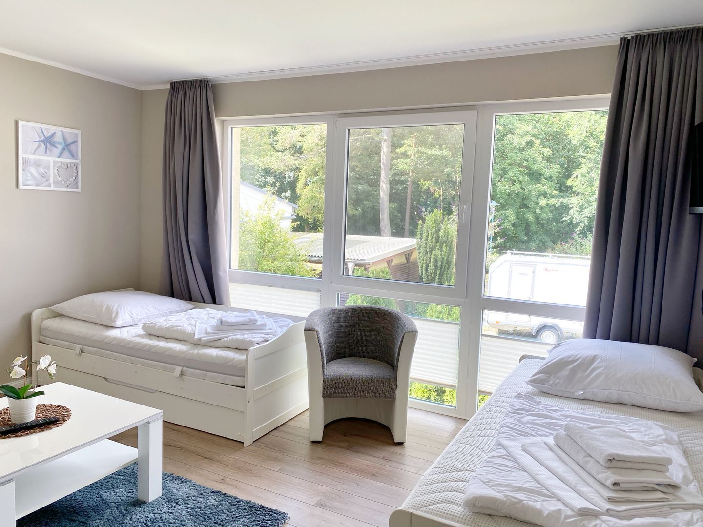 Ein Schlafzimmer mit zwei Betten, einem Sessel, einem Tisch und einem großen Fenster mit Blick auf Grünflächen und ein Gebäude.