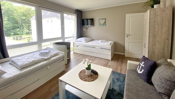 Ein modernes, helles Zimmer mit großem Fenster, einem Bett, einer Couch und einem kleinen Tisch mit Pflanze.