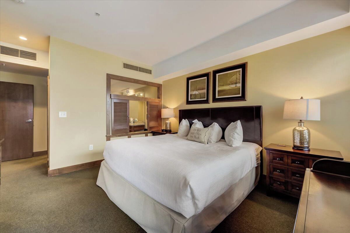 A hotel room with a large bed, a nightstand with a lamp, a dresser, a mirror, and two framed pictures on the wall.