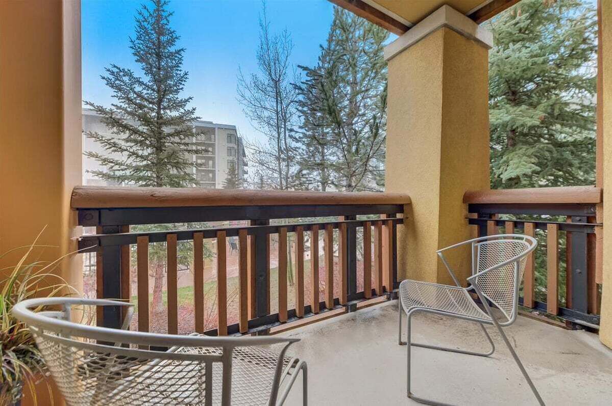 A balcony with two metal chairs, a potted plant, and a view of trees and buildings.