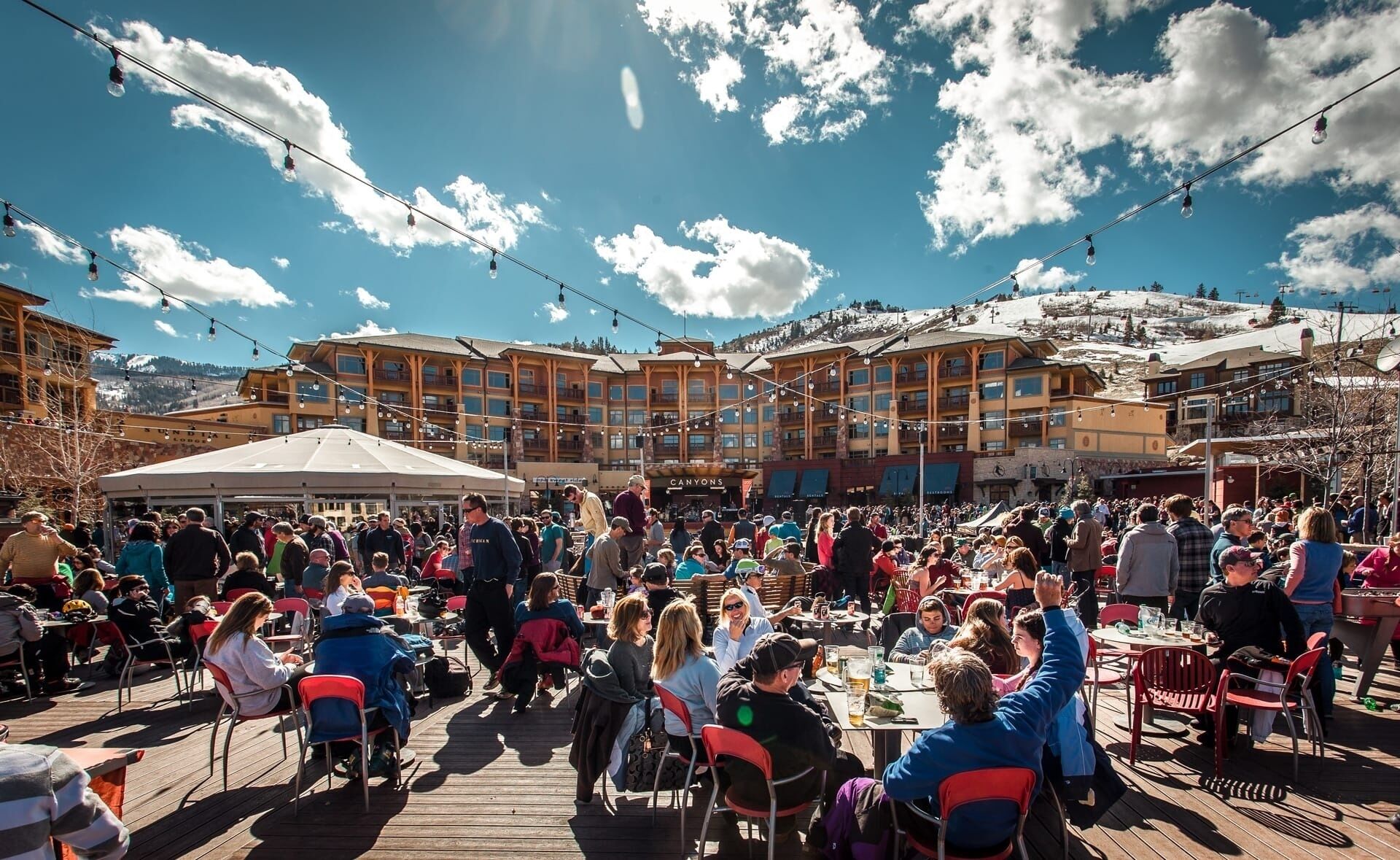 Un espace extérieur animé, aménagé pour les repas, avec des tables et des chaises, un grand bâtiment doté de nombreux balcons et un ciel bleu dégagé parsemé de nuages.