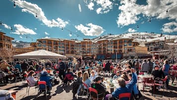 Un espace extérieur animé, aménagé pour les repas, avec des tables et des chaises, un grand bâtiment doté de nombreux balcons et un ciel bleu dégagé parsemé de nuages.