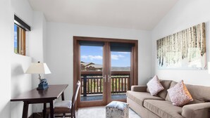 Una habitación con un escritorio de madera, una silla y un sofá color beige con cojines estampados. Un pequeño otoman blanco se encuentra frente al sofá. Se ve un ventanal con vistas a edificios y colinas.