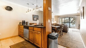 Cocina moderna con armarios de madera, lavavajillas integrado y una estación de café. Hay un comedor con mesa y sillas, y vistas al exterior a través de amplios ventanales.