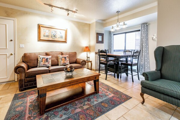 A living room with a brown leather sofa, a wooden coffee table, and a patterned rug. There is a dining area with a table and chairs, and a green armchair. - Shadow Ridge 204-3 | Walk to Lifts | Kitchen | Fireplace | Pool & Hot Tub | Sauna | Fitness Room (Park City)