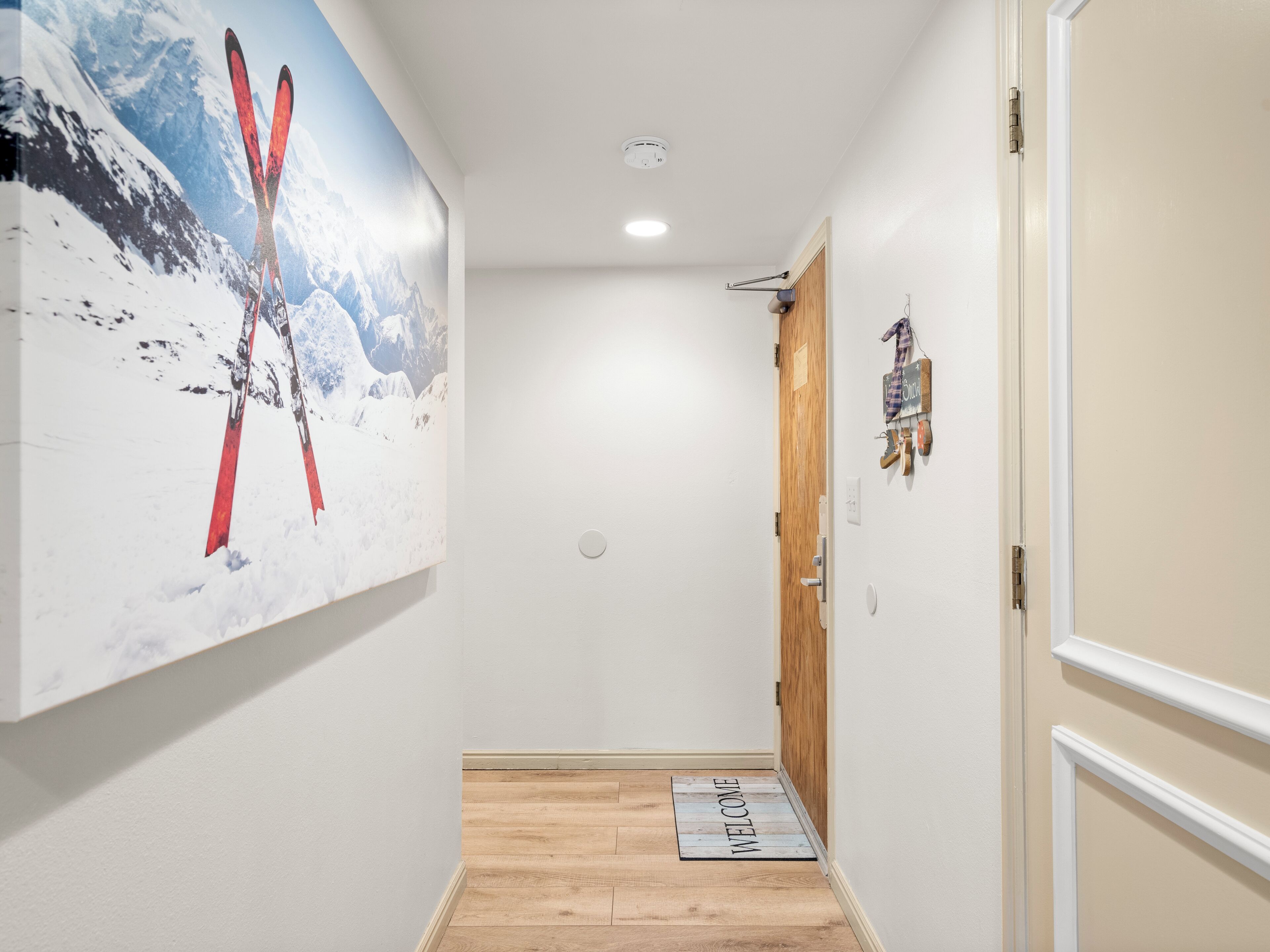 A hallway with a wooden floor, a welcome mat, a door with a sign, and a wall with a large ski-themed painting.
