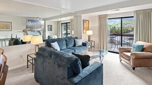 A spacious living room with a large blue sofa, a smaller beige sofa, and a glass coffee table. There is a fireplace with a mantel, a painting, and a view of a balcony with a railing.