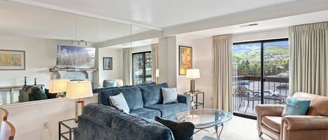 A spacious living room with a large blue sofa, a smaller beige sofa, and a glass coffee table. There is a fireplace with a mantel, a painting, and a view of a balcony with a railing.