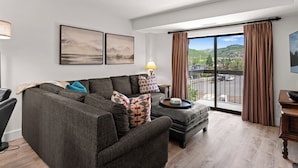 A modern living room with a sectional sofa, a small table with a lamp, and a view of a mountainous landscape through large windows. - Shadow Ridge 406-3 | Walk to Lifts | Mtn Views | Kitchen | Fireplace | Updated | Pool & Hot Tub (Park City)