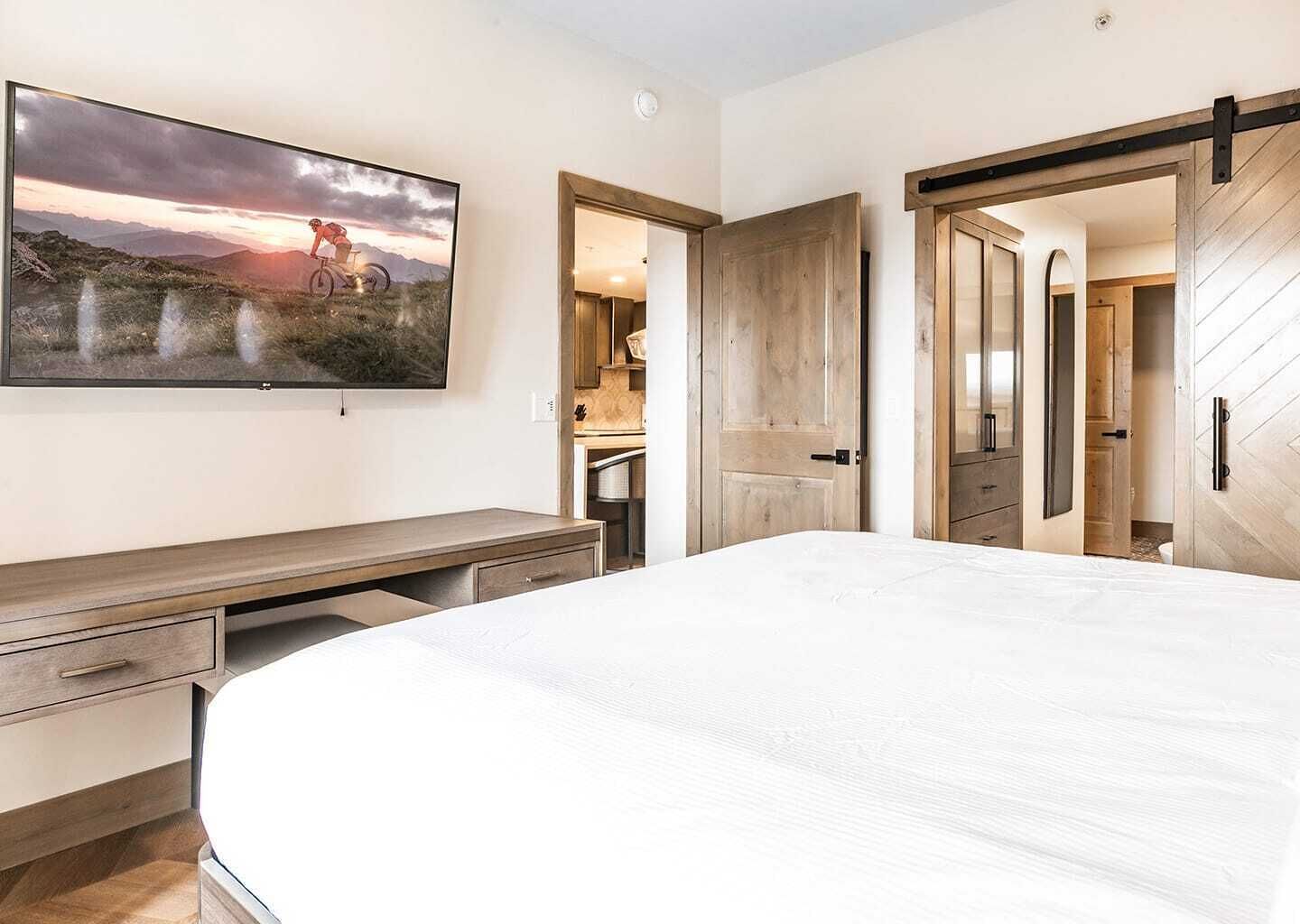Un dormitorio con una cama grande, un escritorio con cajones, un televisor montado que muestra un paisaje, y puertas de madera que dan a otras habitaciones.