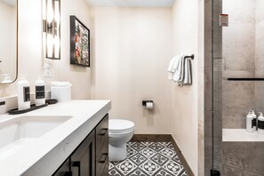 Une salle de bain moderne avec un lavabo, des toilettes et une douche, le tout de couleur blanche. On trouve des articles de toilette sur le lavabo et un porte-serviettes avec des serviettes.