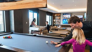 A pool table in a modern room with a large window, a foosball table, and a seating area with couches and a TV. - LIFT 307-A | Ski-In/Out | Near Pool Deck | Mountain View | Hot Tub | Pool | Great Amenities (Park City)