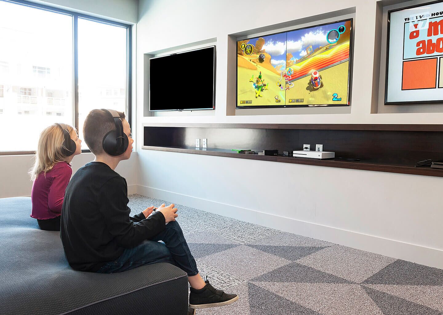 Two people sitting on a bench, watching a video game on a wall-mounted screen.