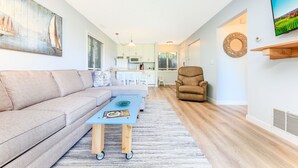 Una sala de estar moderna con un sofá, una mesa de centro y una silla. Se ve una cocina al fondo con gabinetes blancos y un microondas.
