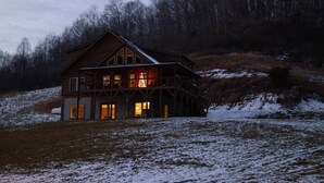 A large log cabin with multiple windows and a balcony, surrounded by a snowy landscape and forested hills.