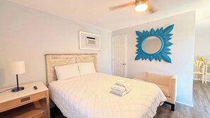 A bedroom with a bed, a nightstand, a lamp, a chair, a door, and a wall-mounted mirror.