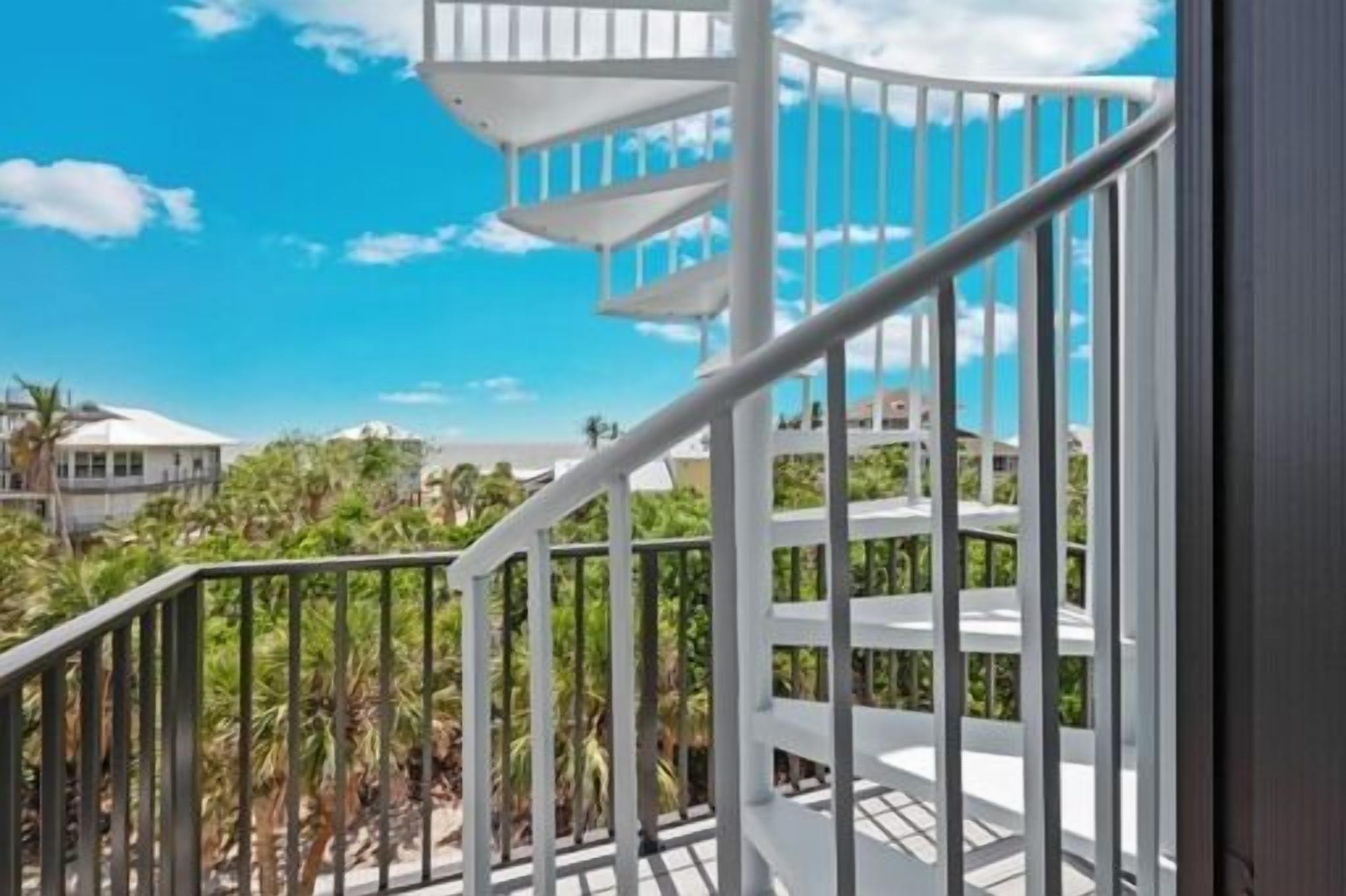 A spiral staircase with white railings leading to a balcony with a clear view of palm trees and buildings.