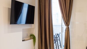 Un salón moderno con un televisor de pantalla plana montado en la pared, un estante con una planta y vistas al exterior a través de amplias ventanas con cortinas marrones.