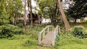 Eine Holzbrücke führt zu einem Haus, umgeben von üppigem Grün.