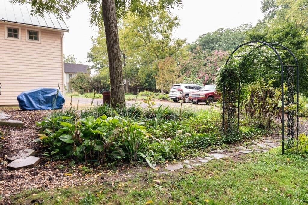 Un jardín con un arco de metal, un objeto cubierto y una casa al fondo.