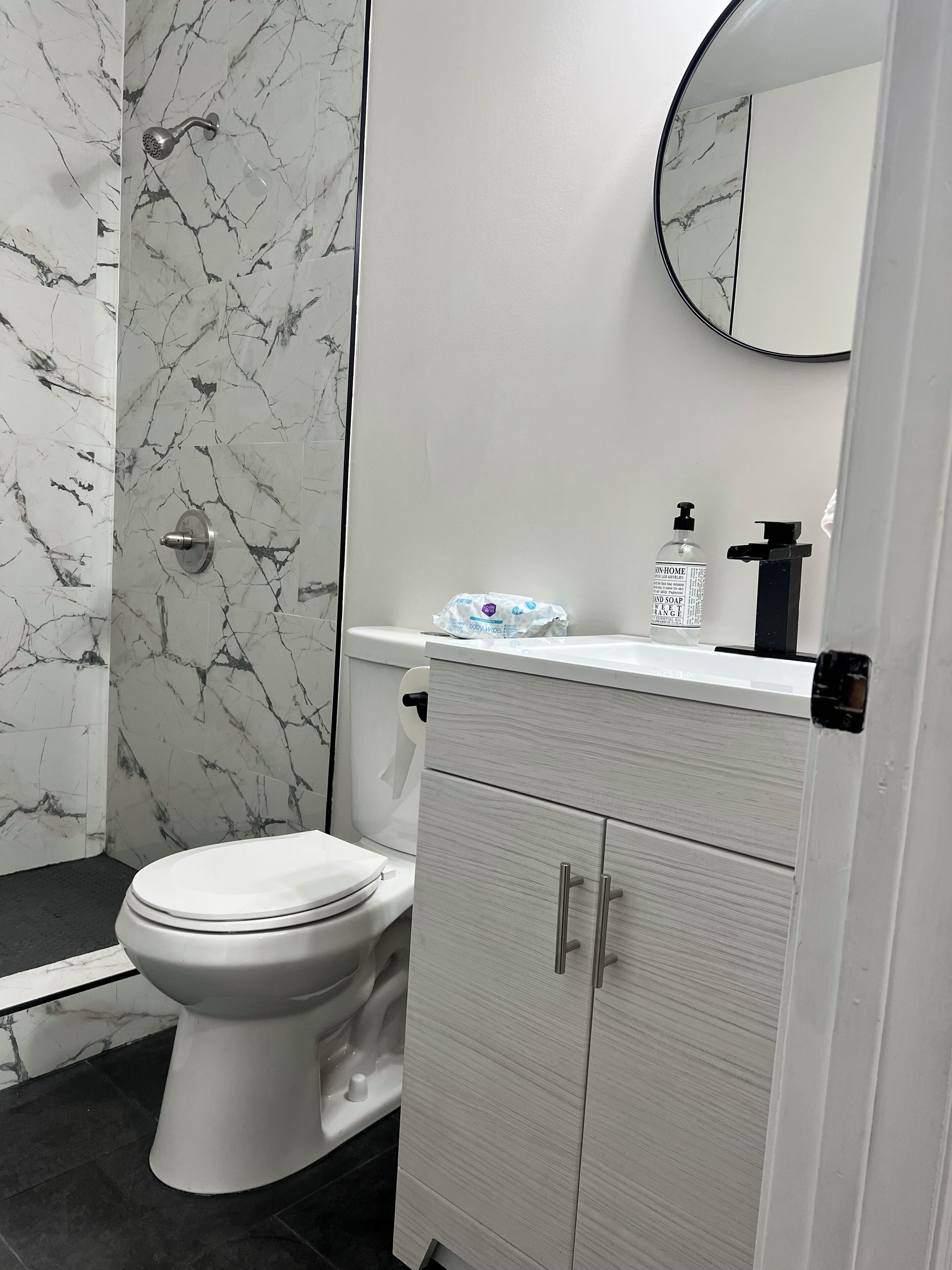 Ein Badezimmer mit Toilette, einer Dusche mit Marmorwand, einem Waschbecken mit Spiegel und einem Schrank mit Schubladen.