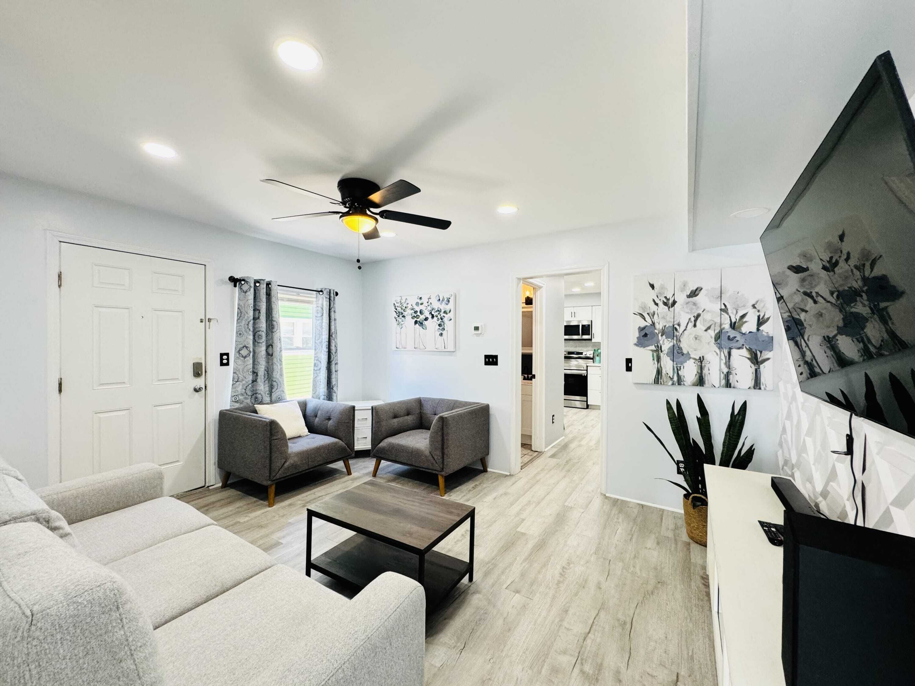 A modern living room with a ceiling fan, grey sofas, a coffee table, and a kitchen in the background.