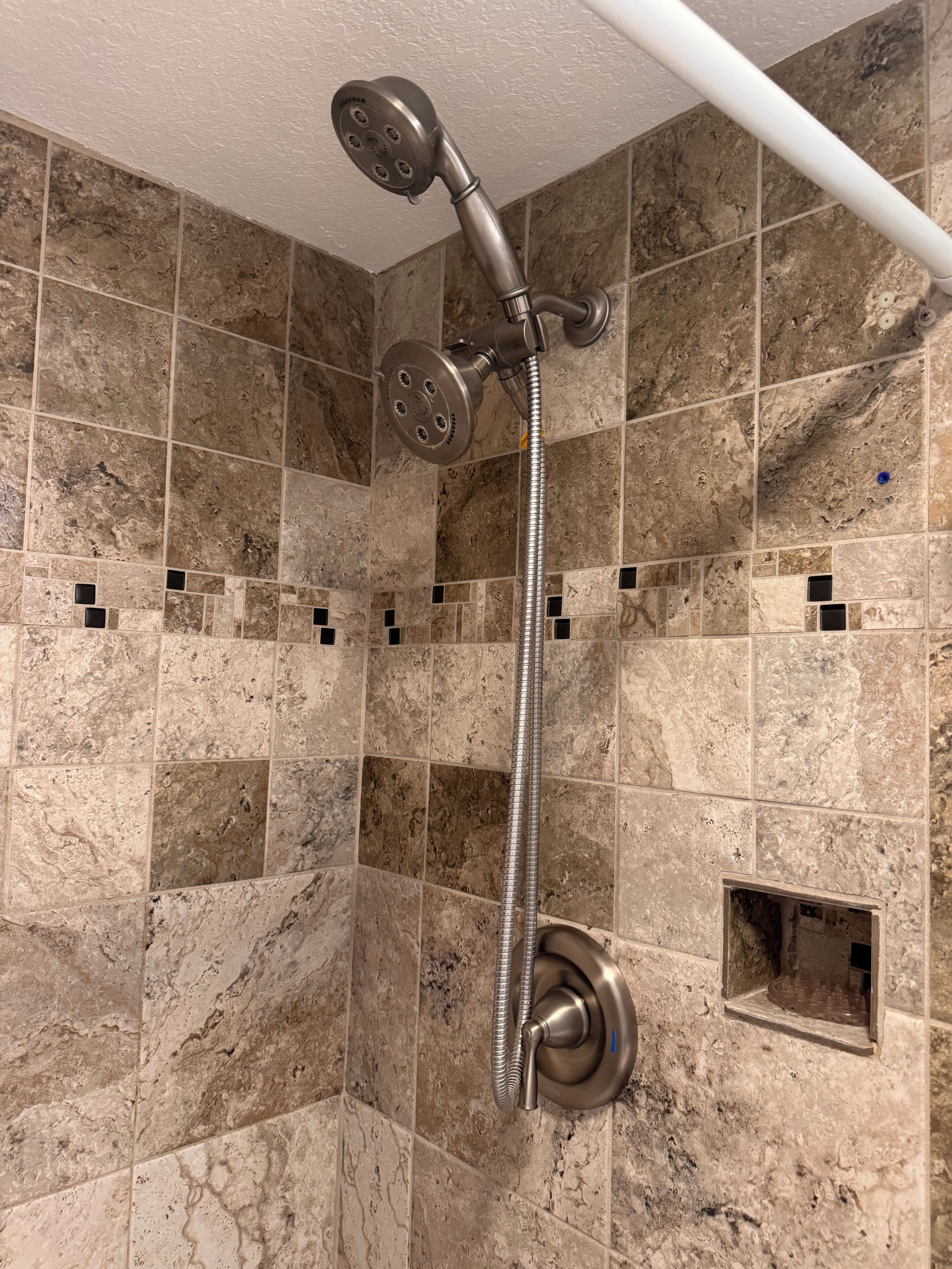 Eine Dusche mit Handbrause und flexiblem Duschschlauch, vor einer Wand mit quadratischen Fliesen und einem kleinen, quadratischen Lüftungsgitter.