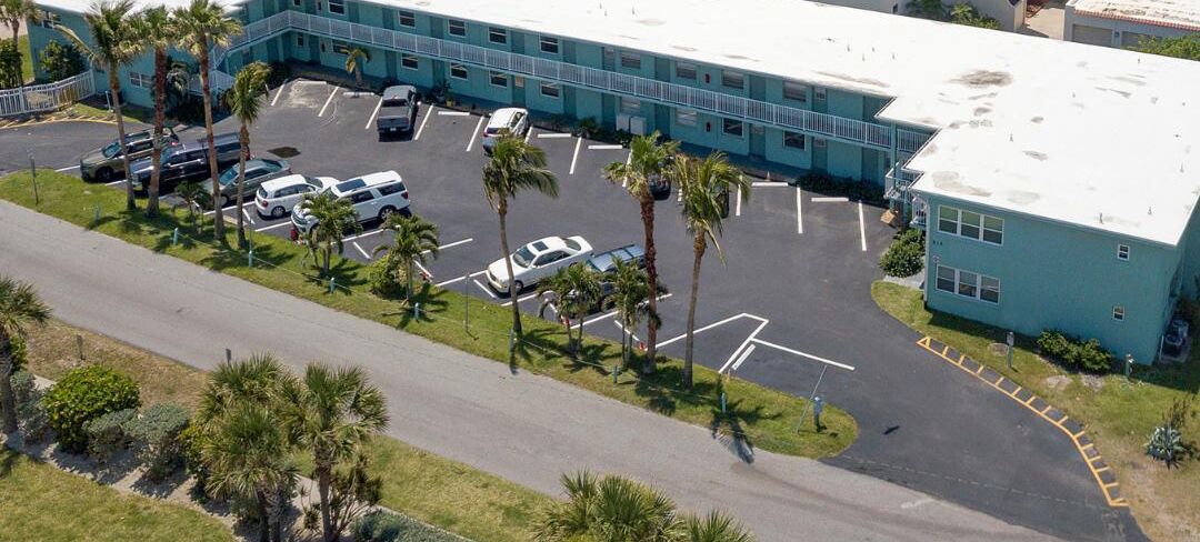 Vista aérea de un hotel con estacionamiento, palmeras y un edificio de fachada azul claro.