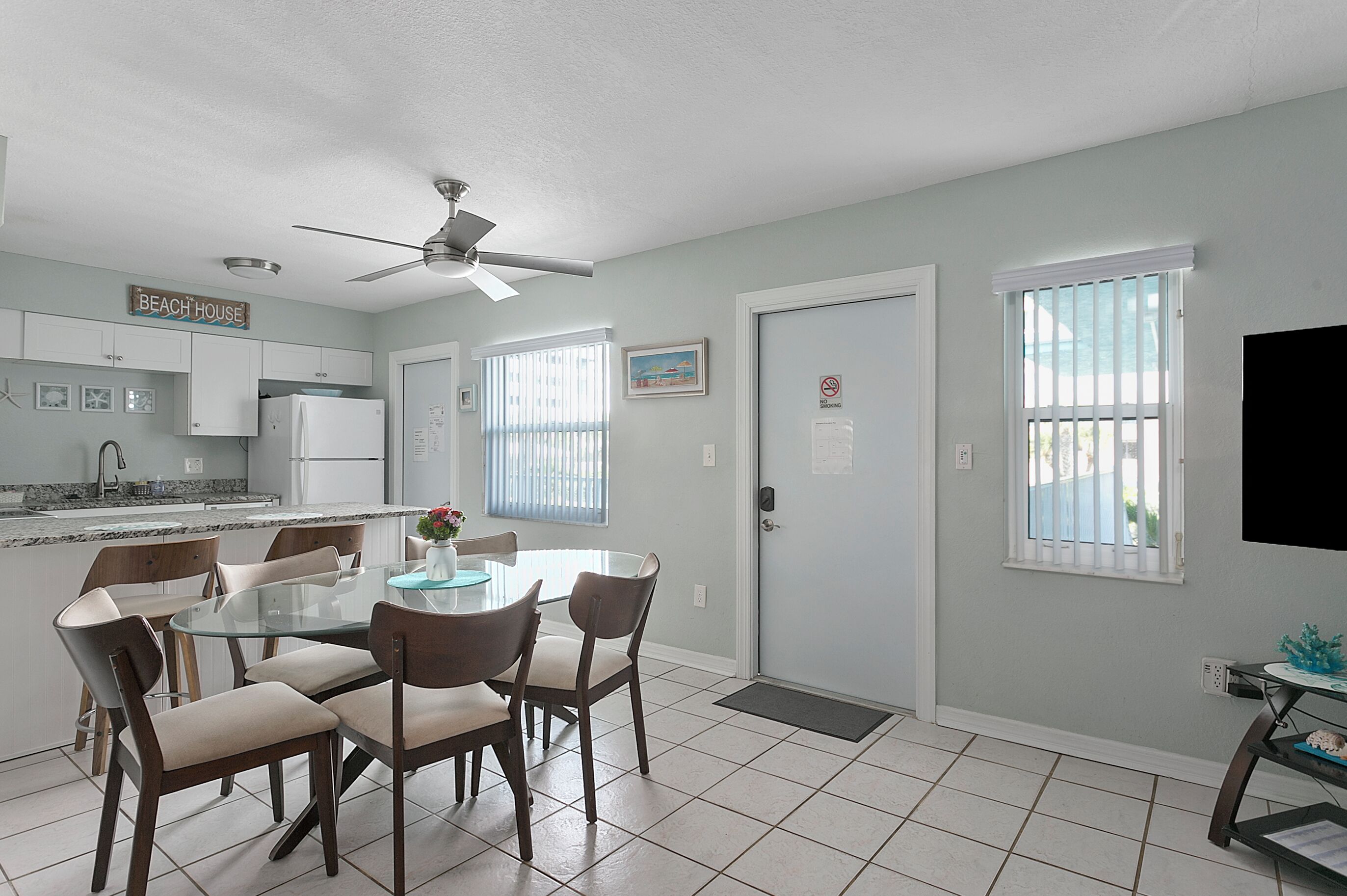 A modern kitchen with white cabinets, a dining area with a glass table and chairs, and a ceiling fan.