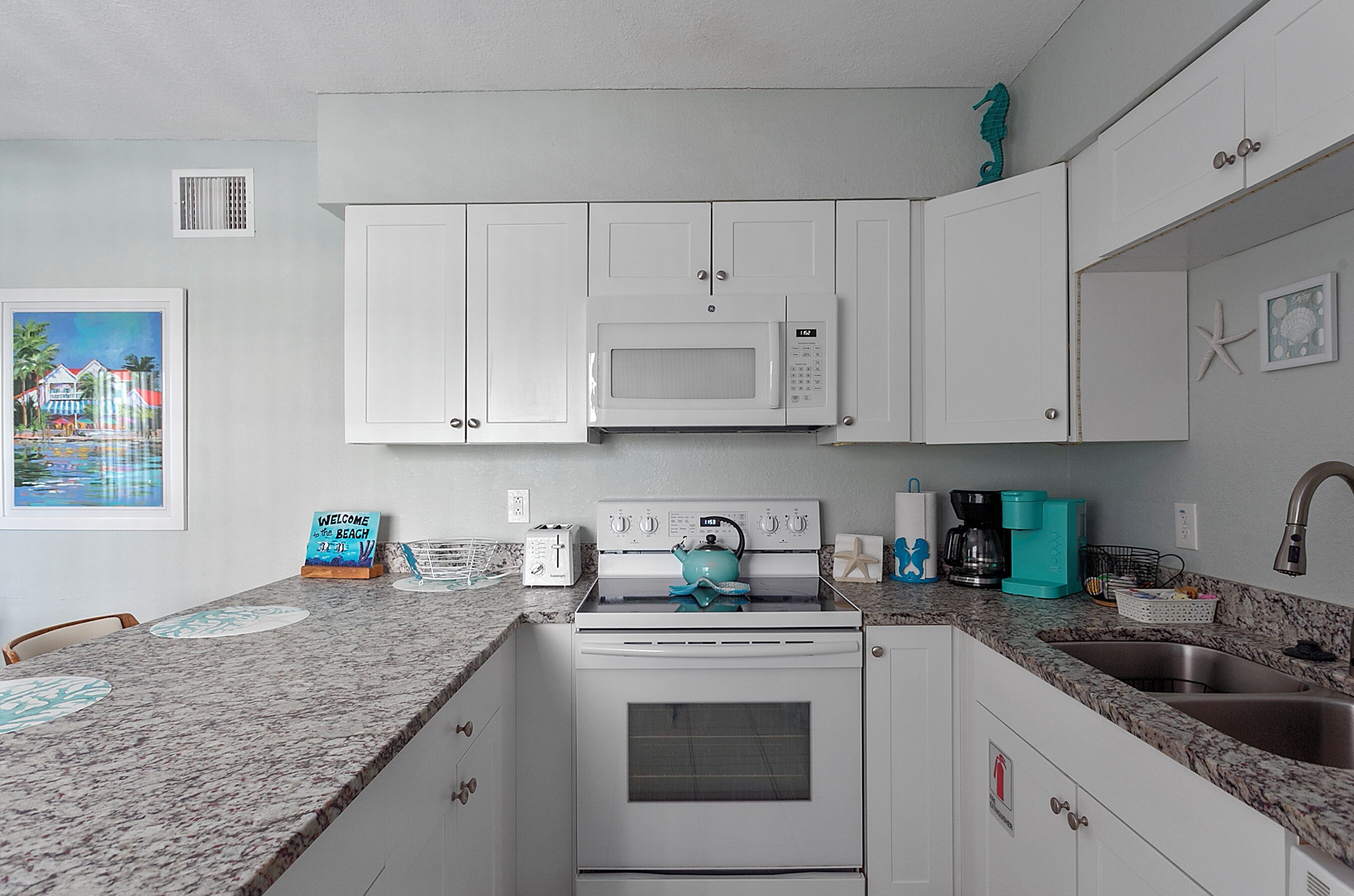 Una cocina compacta con gabinetes blancos, microondas, estufa y lavamanos. Hay un cuadro en la pared y un cartel de bienvenida.