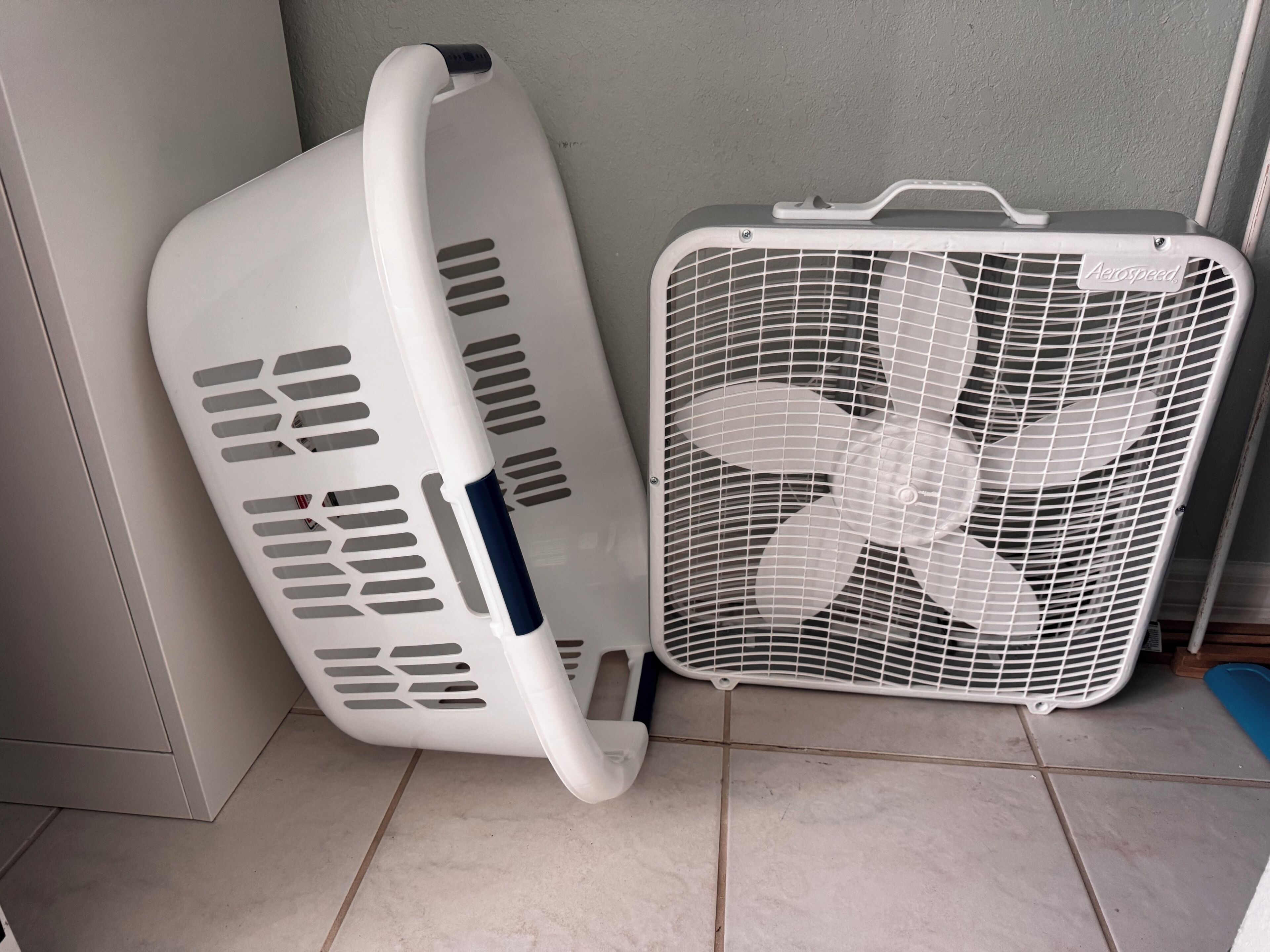 A corner of a room with a white laundry basket leaning against a wall and a white fan with a grid pattern.